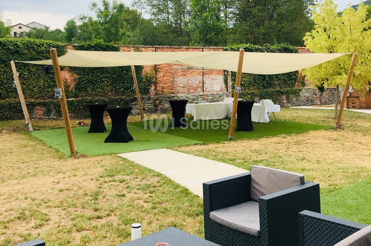Espace extérieur aménagé avec tables hautes, chaises et voiles d'ombrage, entouré de verdure et d'un mur en briques.