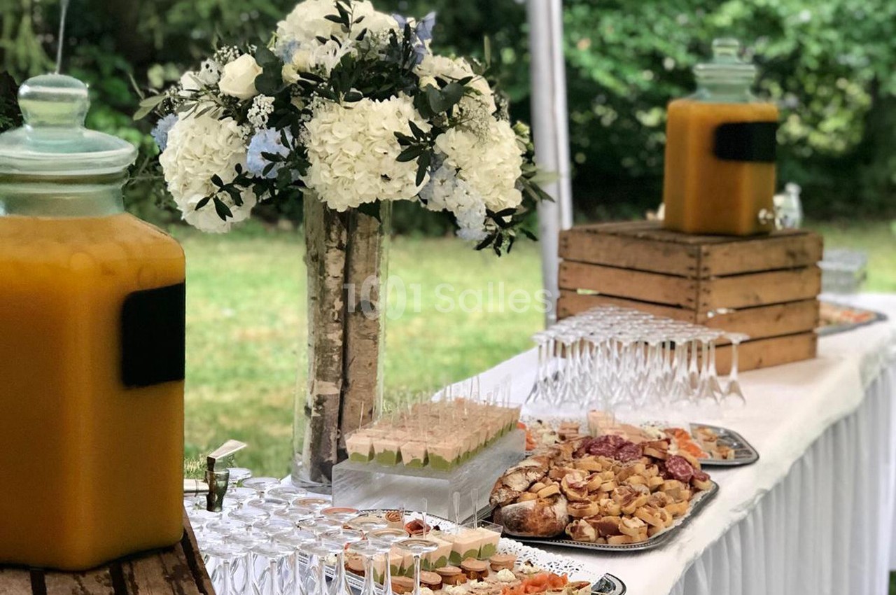 Buffet en extérieur avec boissons, fleurs blanches, verrines, charcuterie et amuse-bouches sur une table blanche.