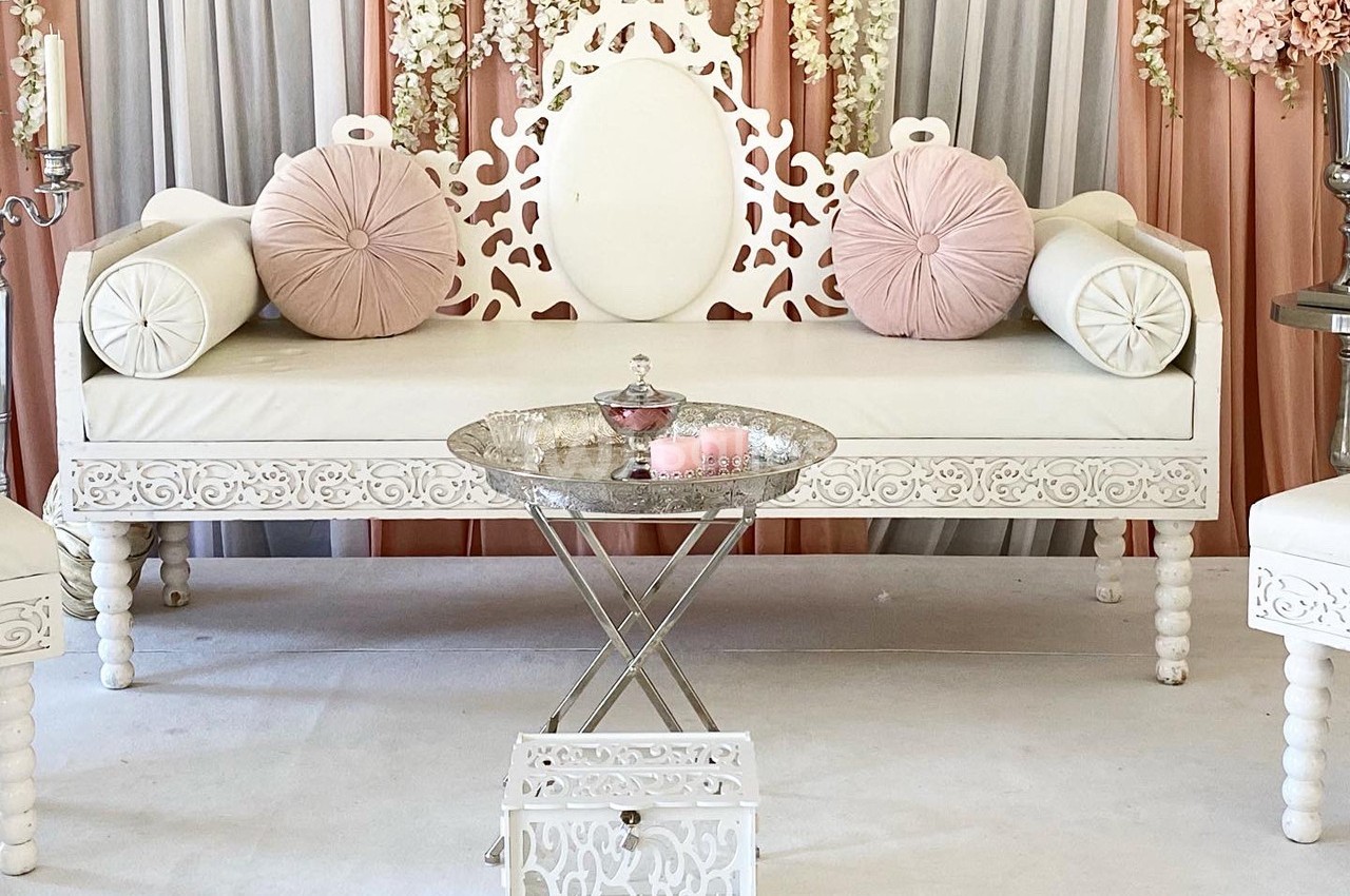 Canapé blanc sculpté avec coussins roses, table basse argentée et décor floral en arrière-plan.