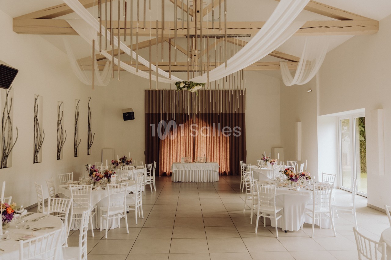Salle de réception lumineuse avec tables décorées de fleurs, chaises blanches et plafond orné de voilages.