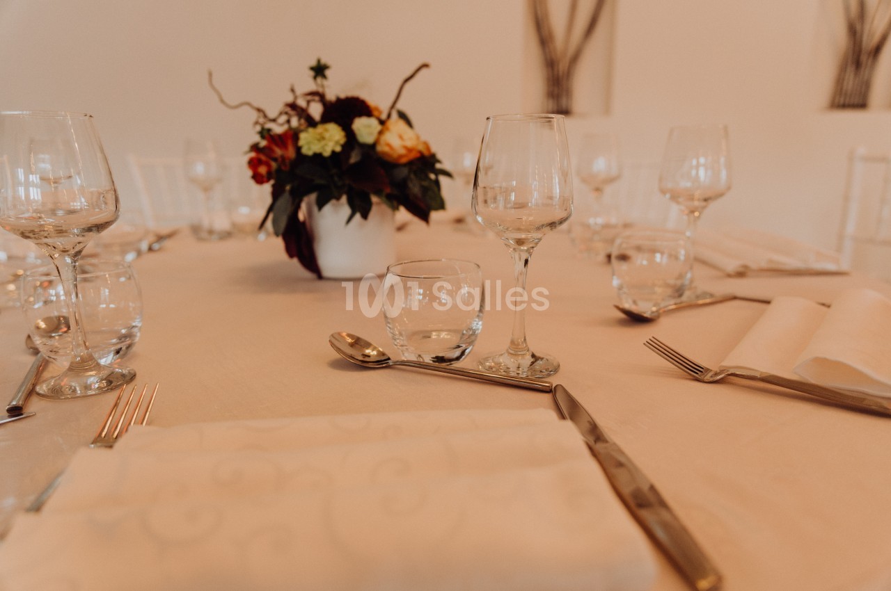 Table dressée avec nappes blanches, verres, couverts et un centre de table floral dans un décor épuré.