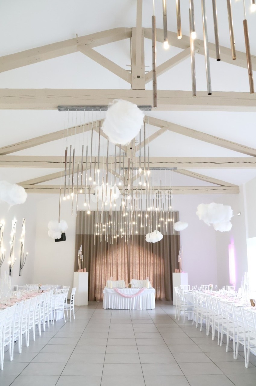 Salle de réception lumineuse avec tables blanches alignées, décorations suspendues et poutres apparentes en bois clair.