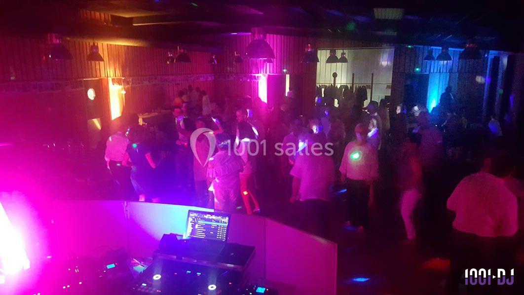 Des personnes dansent dans une salle faiblement éclairée avec des lumières colorées et une table de mixage au premier plan.