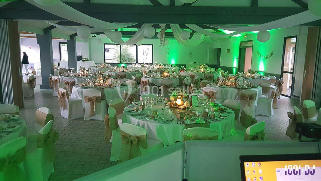 Salle de réception décorée avec des tables rondes, nappes blanches, chaises ornées de nœuds beige et éclairage vert.