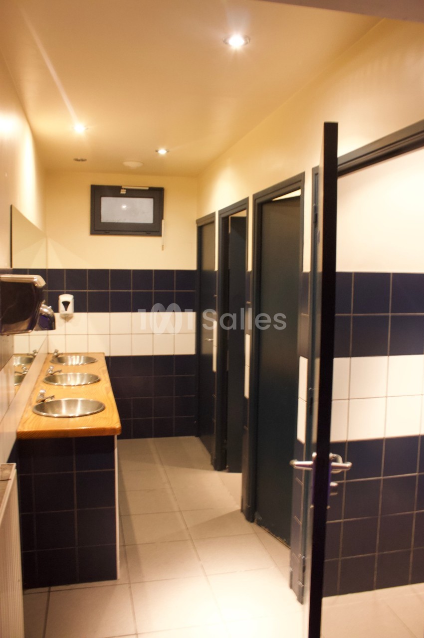 Salle de bain publique avec lavabos en inox, carrelage bleu et blanc, et portes de cabines fermées.