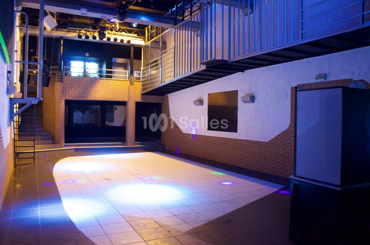 Salle vide avec piste de danse éclairée, murs en briques et métal, et mezzanine au fond.