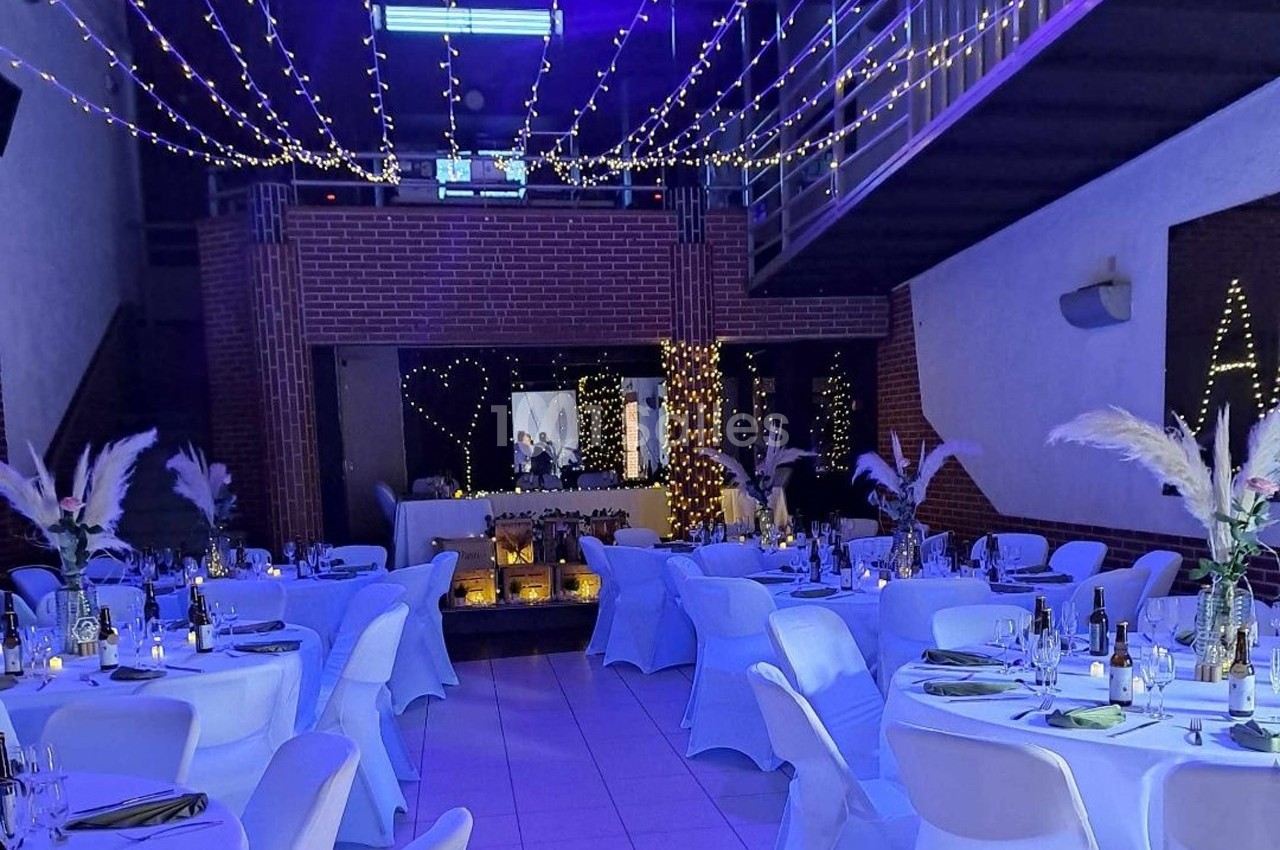 Salle décorée pour un événement, avec tables dressées, guirlandes lumineuses suspendues et ambiance tamisée.