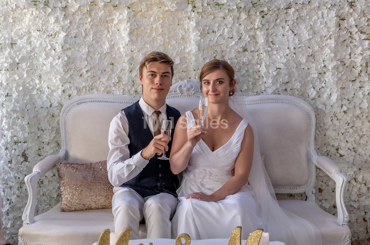 Un couple assis sur un canapé blanc, tenant des flûtes de champagne, devant un mur décoré de fleurs blanches.