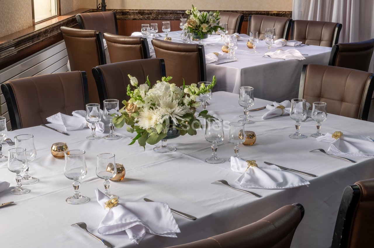 Salle de réception avec tables dressées, nappes blanches, couverts, verres et centre de table floral.