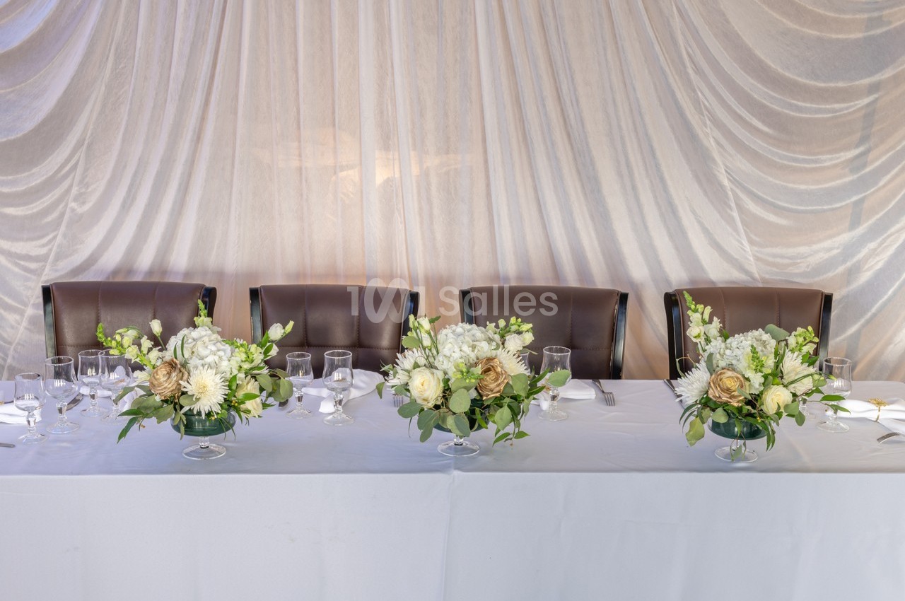 Table décorée avec nappes blanches, chaises en cuir marron et arrangements floraux élégants devant un rideau drapé.
