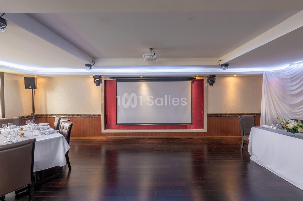 Salle de réception avec parquet sombre, tables dressées et écran de projection encadré par des rideaux rouges.