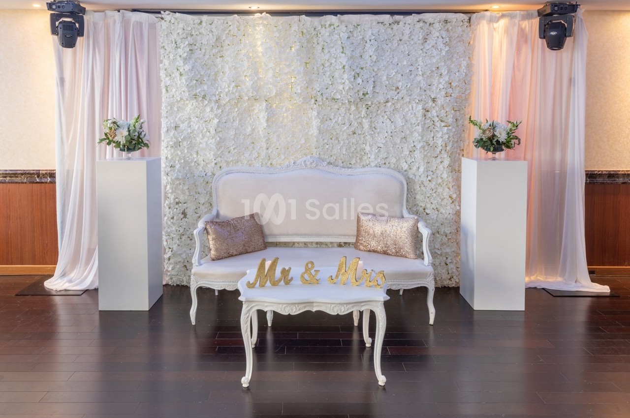 Banc blanc orné de coussins dorés devant un mur de fleurs blanches, encadré de rideaux et de colonnes décoratives.