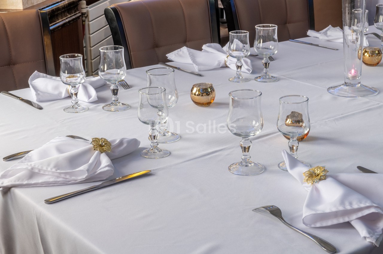 Table dressée avec nappes blanches, serviettes pliées, verres, couverts et petites décorations dorées.