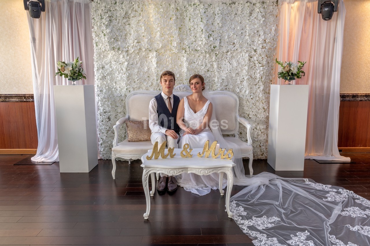 Un couple de mariés assis sur un canapé devant un mur fleuri, avec une table portant l'inscription ’Mr & Mrs’.