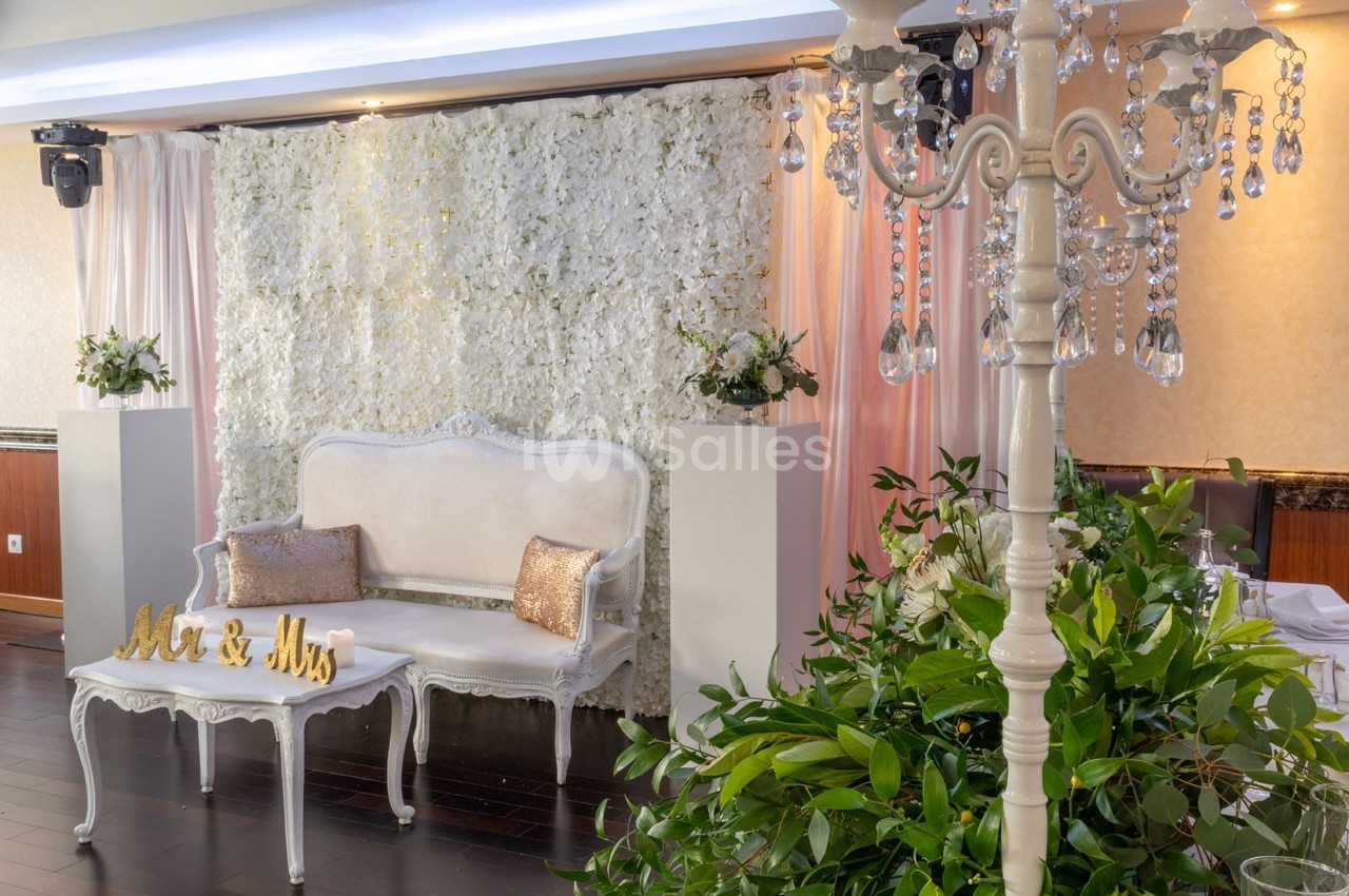 Décor de mariage avec un canapé blanc, un mur de fleurs blanches, des rideaux roses et un chandelier orné de cristaux.