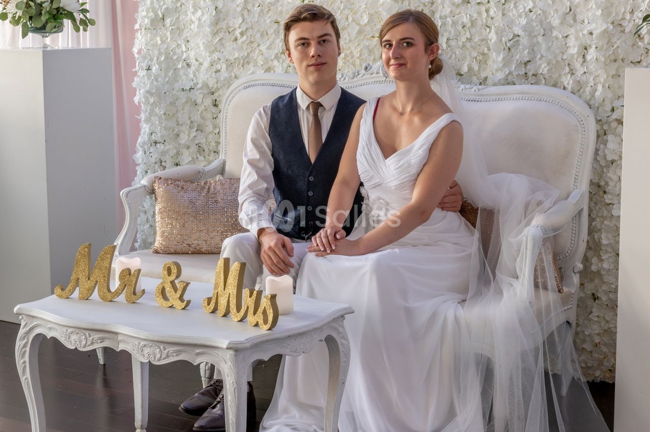 Un couple en tenue de mariage est assis sur un canapé devant un mur de fleurs blanches, avec une décoration ’Mr & Mrs’.