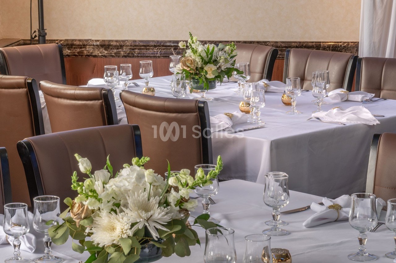 Salle de réception avec tables dressées, nappes blanches, verres, serviettes pliées et bouquets de fleurs.