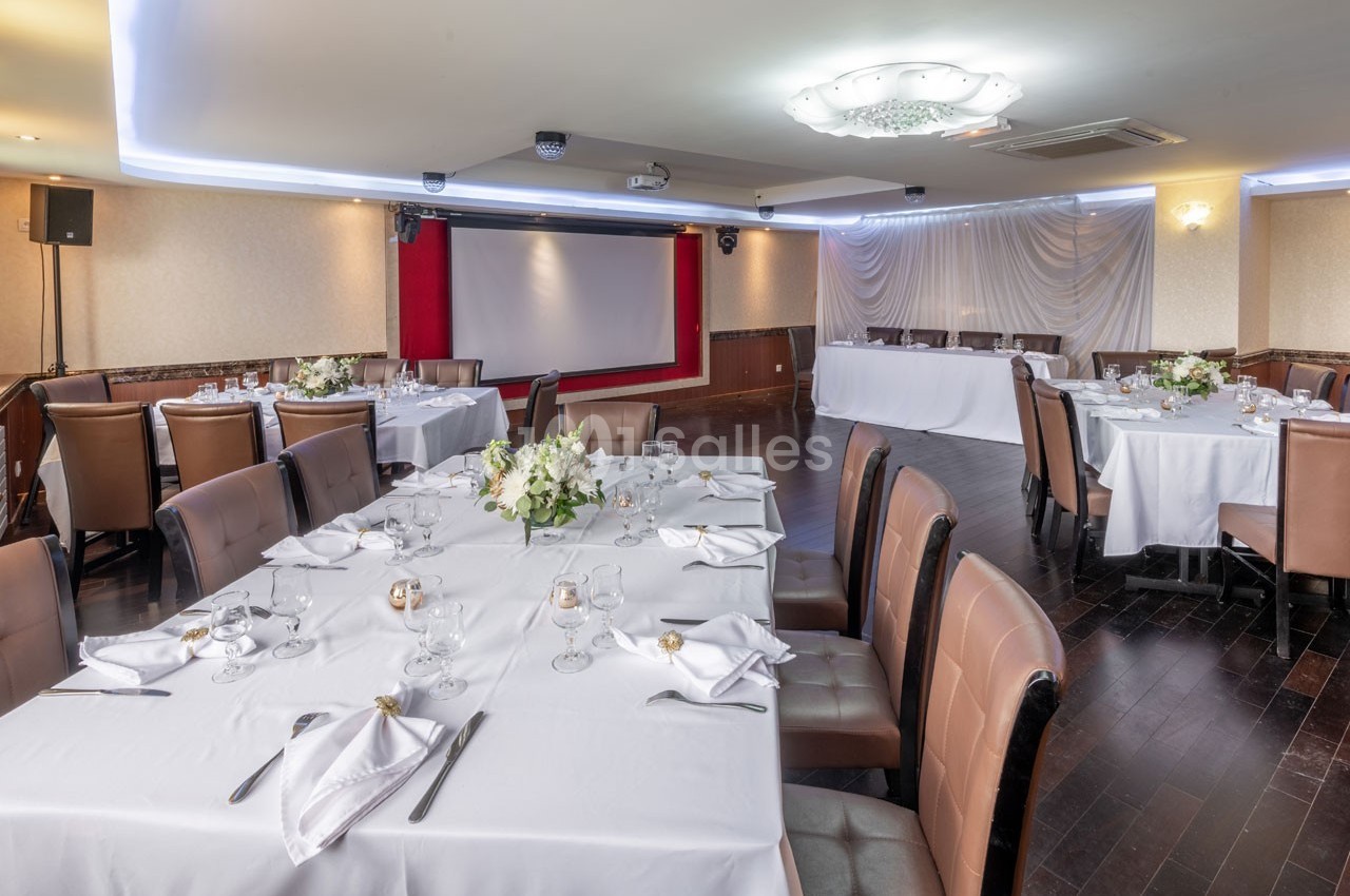 Salle de réception avec tables dressées, nappes blanches, chaises en cuir, décoration florale et écran de projection.