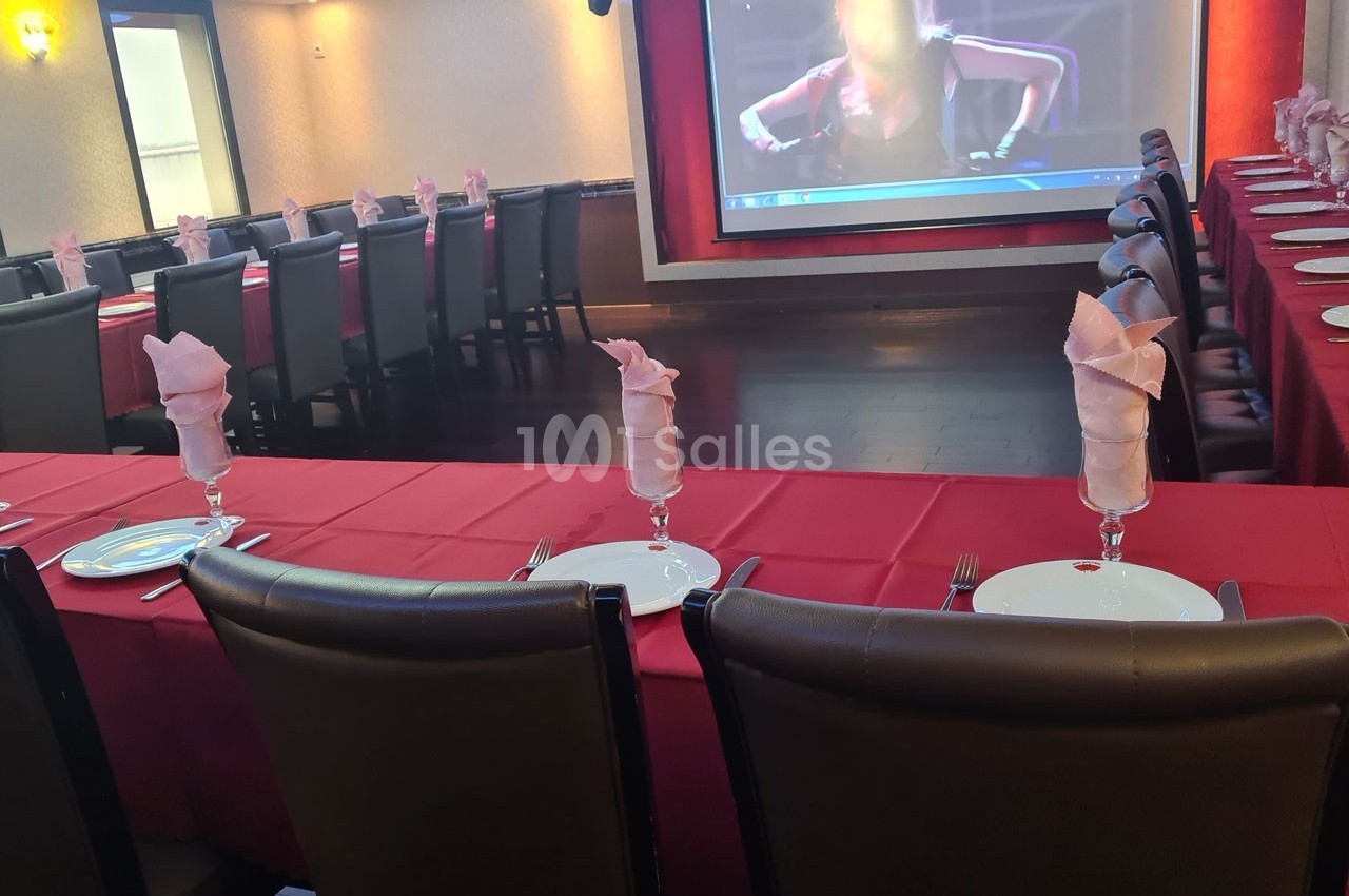 Salle de réception avec tables dressées, nappes rouges, serviettes pliées dans des verres et écran de projection en arrière…
