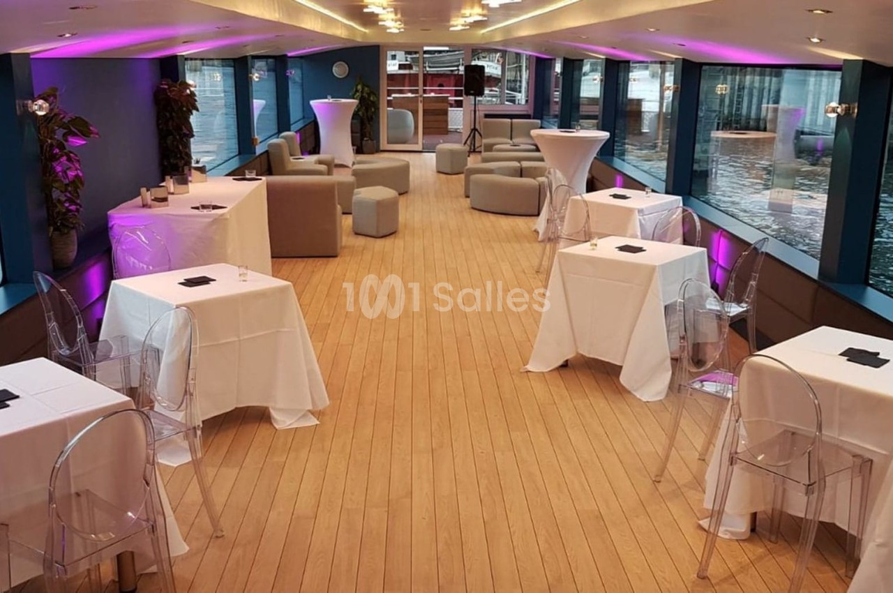 Salle de réception sur un bateau avec tables nappées, chaises transparentes et éclairage rose, vue sur l'eau.