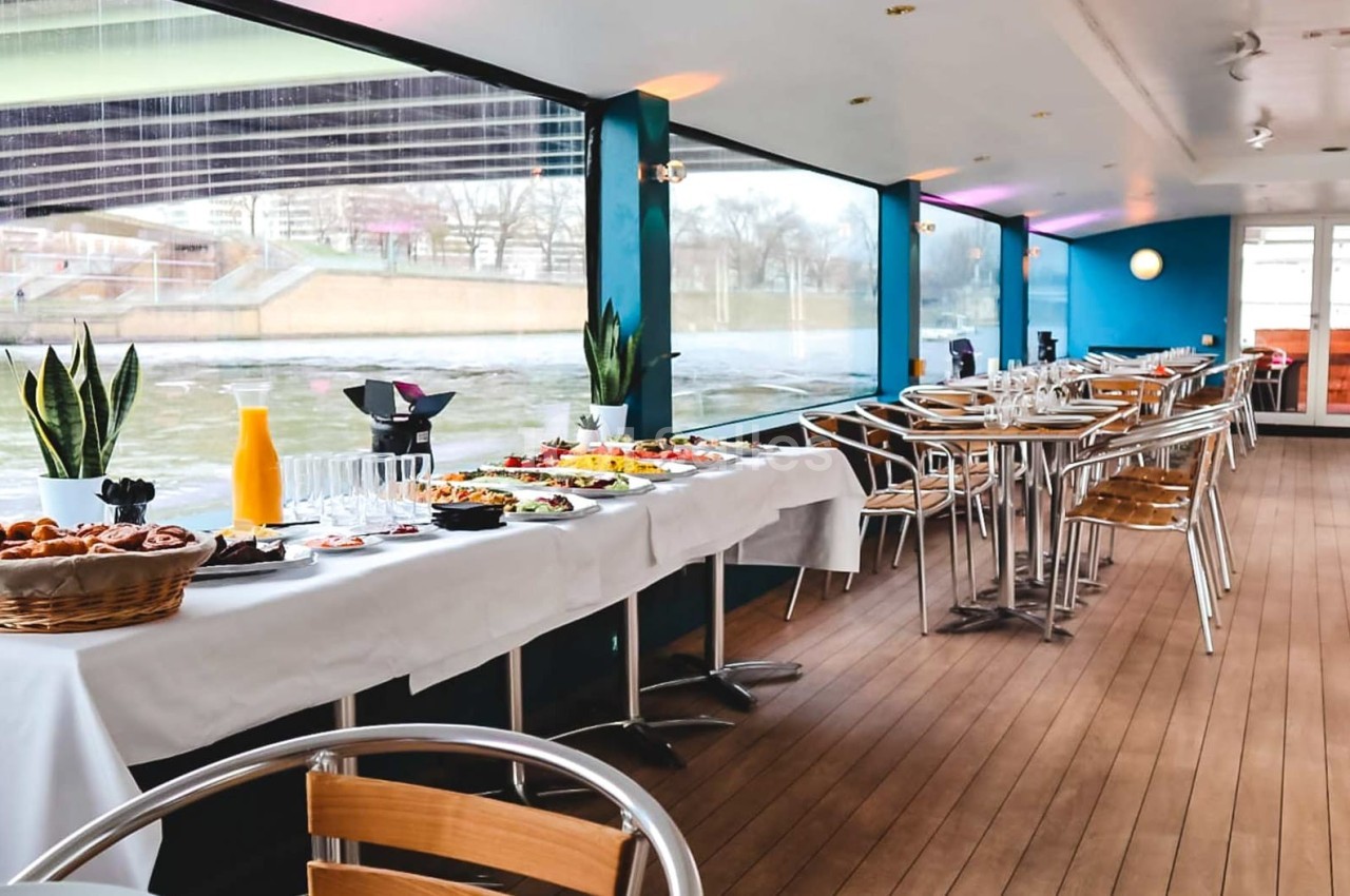 Salle de restaurant sur un bateau avec tables dressées et buffet varié, vue sur l'extérieur à travers de grandes baies…