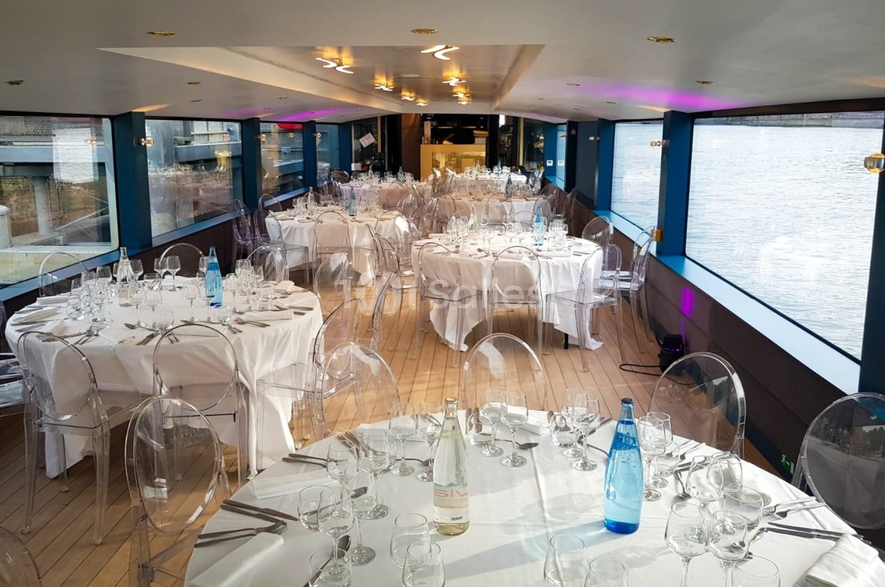 Salle de réception sur un bateau avec tables rondes dressées, nappes blanches et chaises transparentes, vue sur l'eau.
