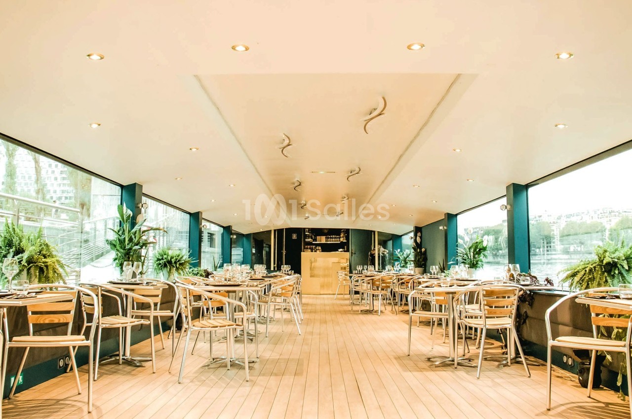 Salle lumineuse avec grandes baies vitrées, tables et chaises en métal, décorée de plantes vertes.
