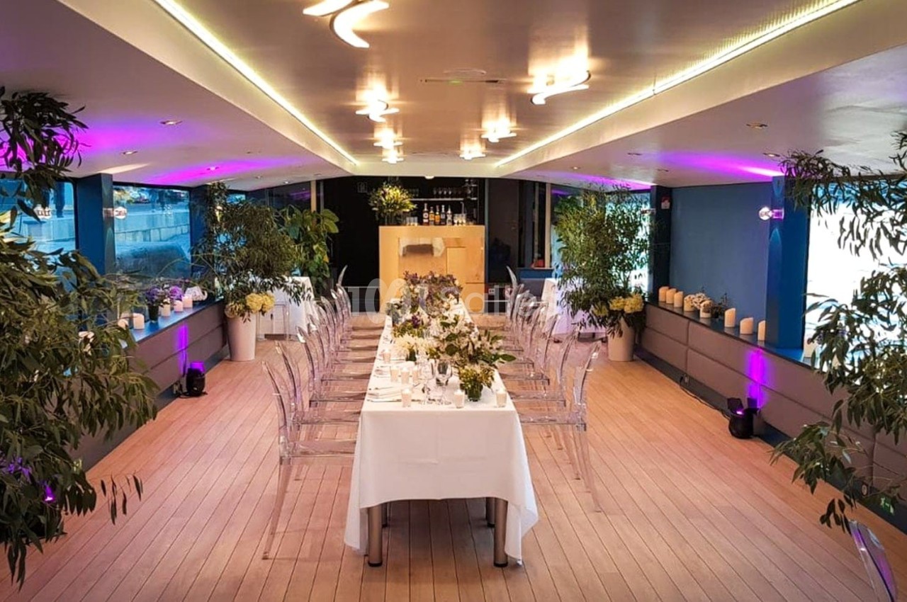 Salle de réception élégante avec tables décorées, chaises transparentes, plantes et éclairage tamisé.
