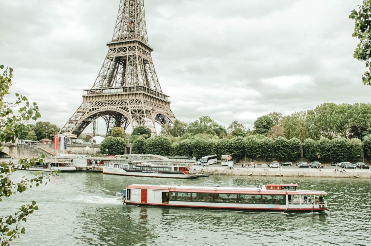 Bateau-mouche naviguant sur la Seine devant la tour Eiffel, entourée d'arbres et d'un ciel nuageux.