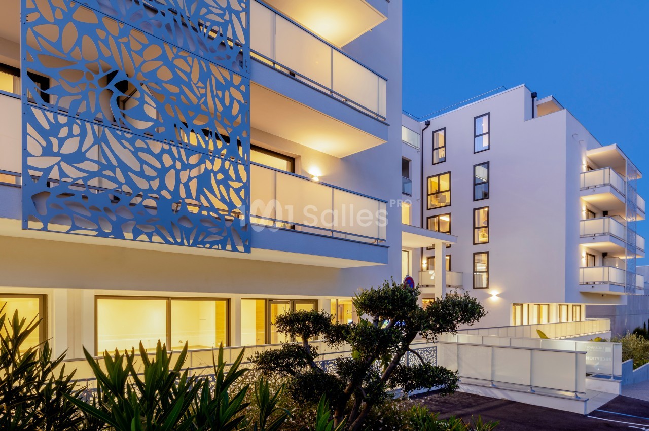 Façade moderne d'un immeuble résidentiel avec balcons éclairés et motifs décoratifs, entouré de végétation.