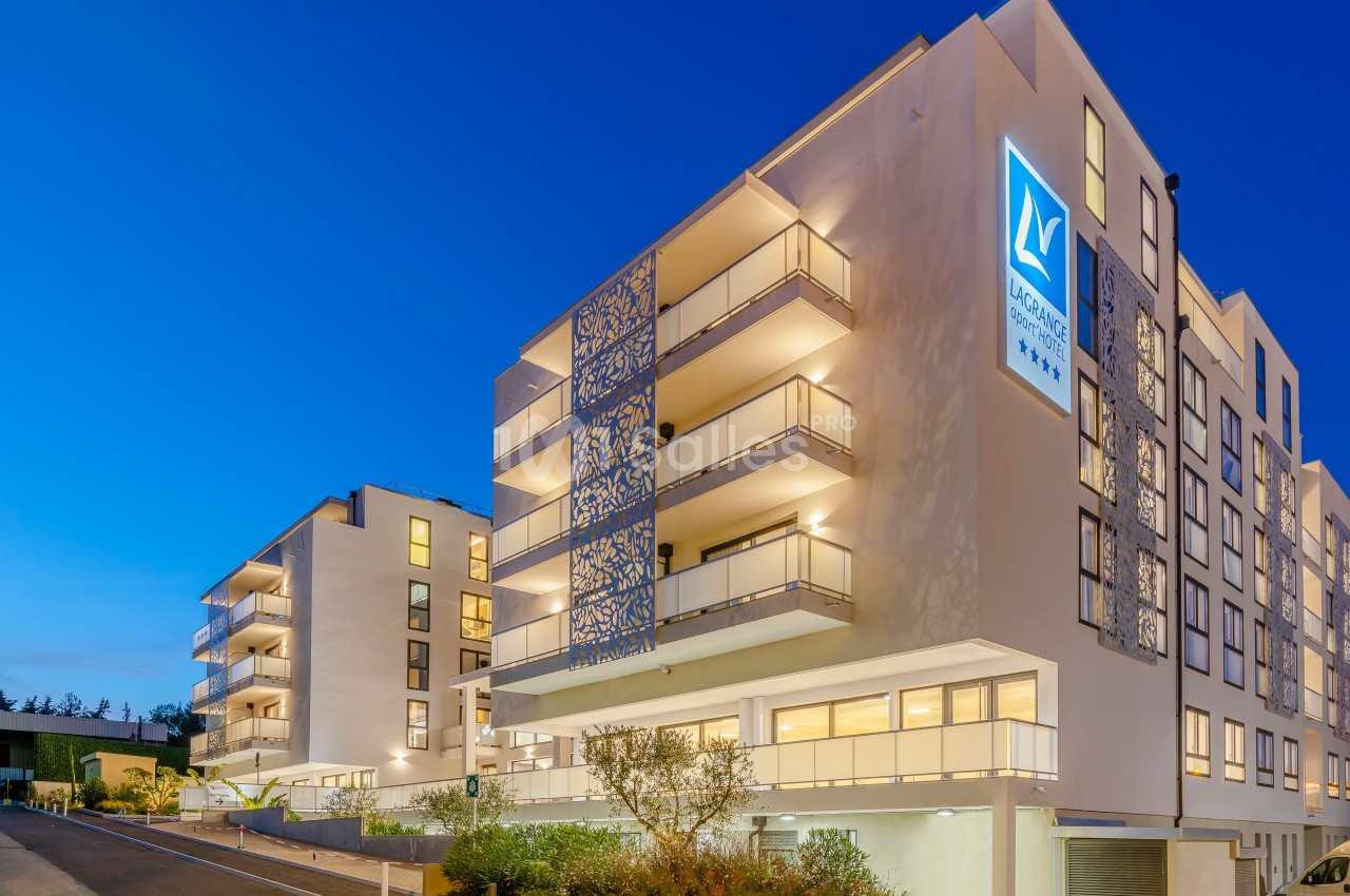 Façade d'un immeuble moderne éclairé en soirée, avec balcons et enseigne lumineuse visible.