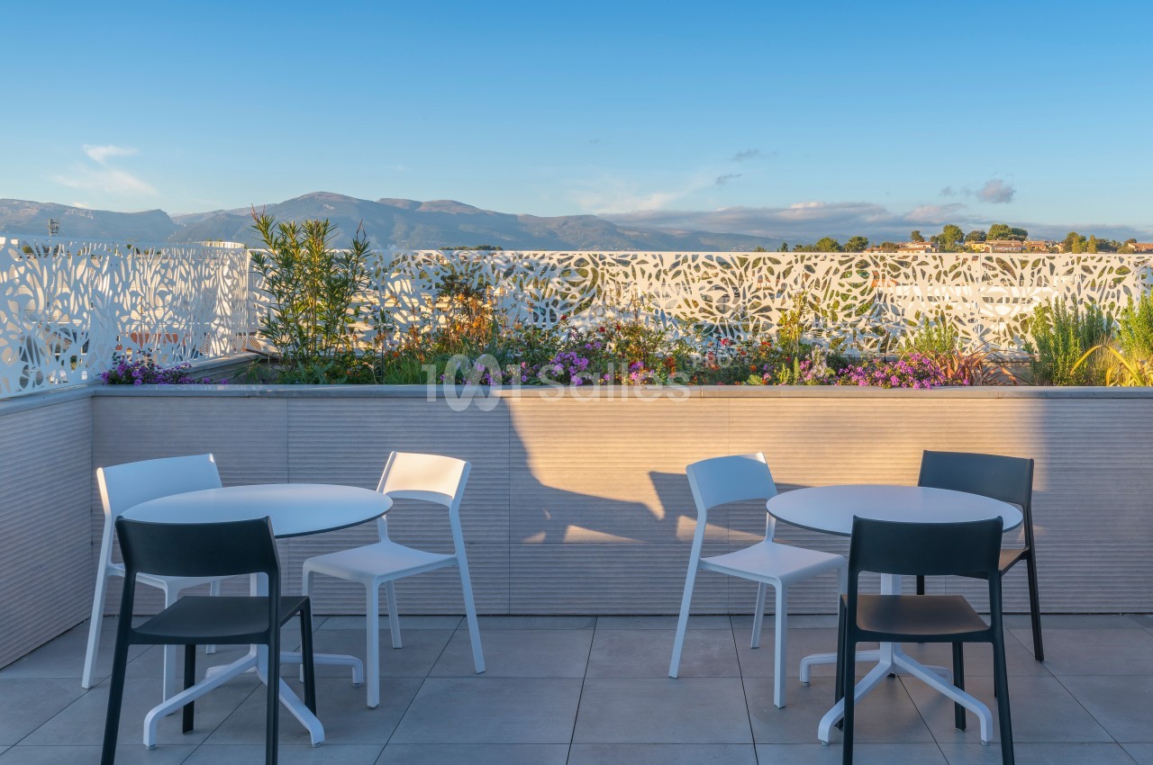 Terrasse avec tables et chaises blanches et noires, entourée de plantes et d'une balustrade ajourée, vue sur des montagnes.