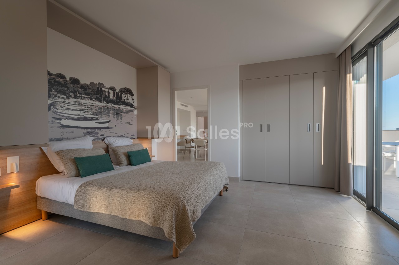 Chambre lumineuse avec lit double, décoration murale représentant un paysage, armoire intégrée et accès à un balcon.
