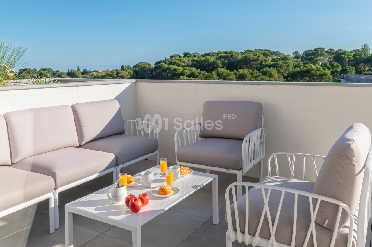 Salon de jardin avec canapés et table basse, petit-déjeuner servi, terrasse ensoleillée avec vue sur des arbres.
