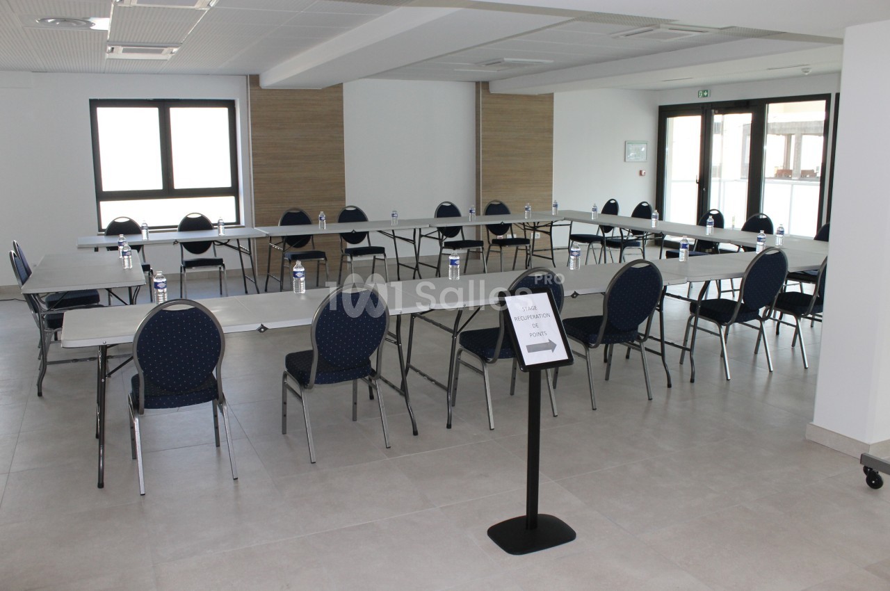 Salle de réunion lumineuse avec tables disposées en U, chaises bleues et bouteilles d'eau sur les tables.