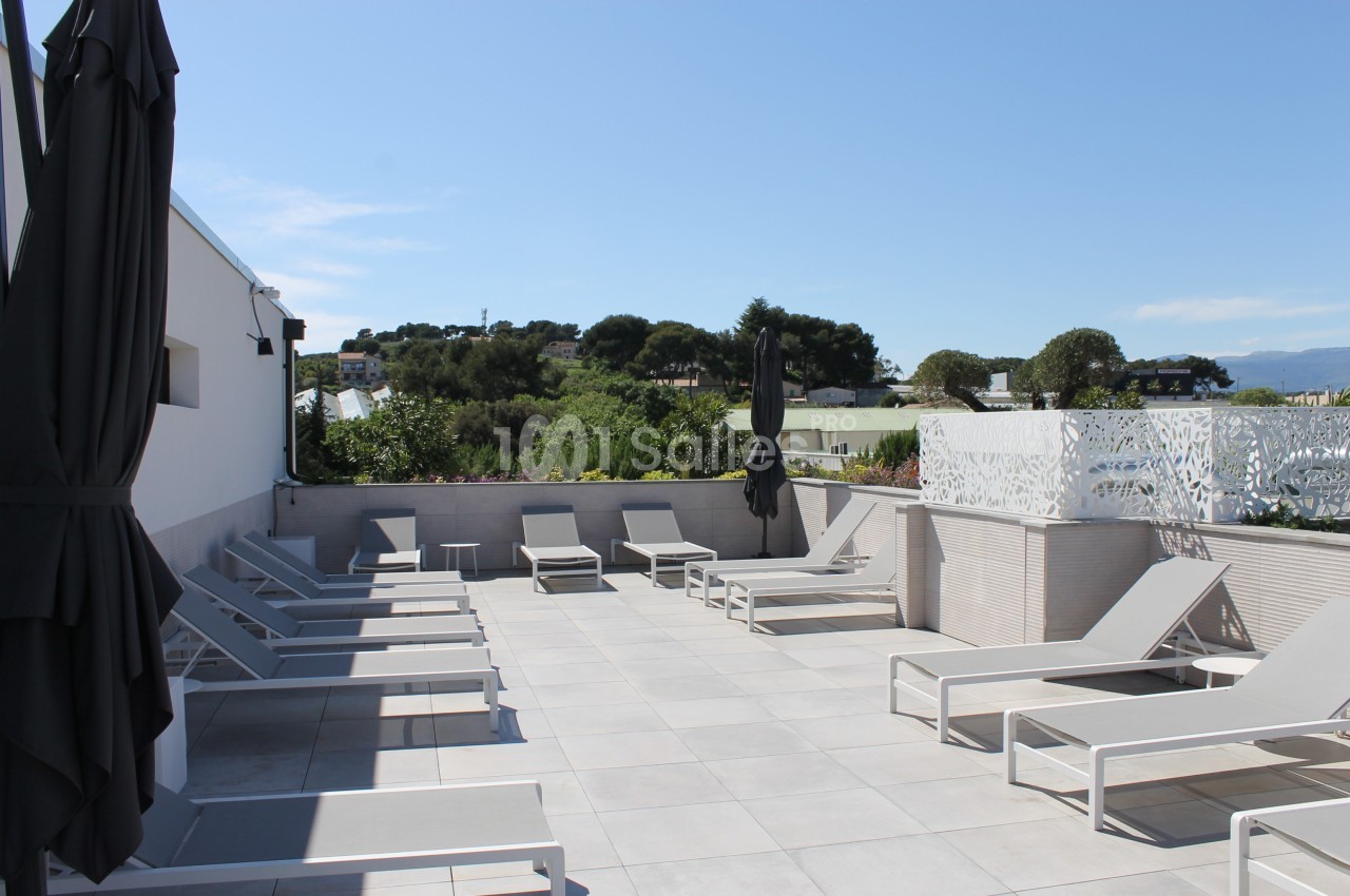 Terrasse ensoleillée avec transats gris, parasols noirs et vue sur un paysage verdoyant.