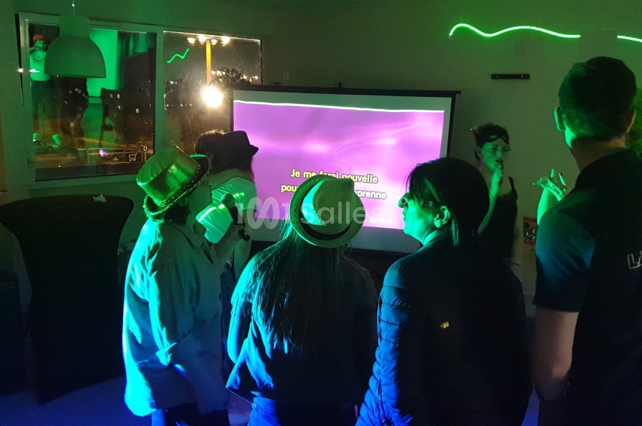 Des personnes chantent devant un écran de karaoké dans une pièce éclairée par des lumières colorées.