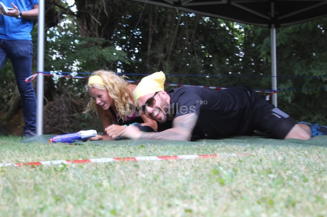 Deux personnes souriantes participent à une activité ludique en extérieur, rampant sur l'herbe sous une structure.