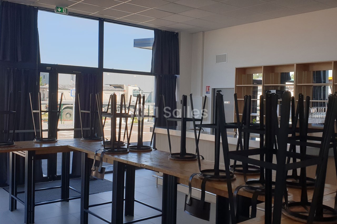 Chaises empilées sur des tables dans une salle lumineuse avec de grandes fenêtres donnant sur un parking.