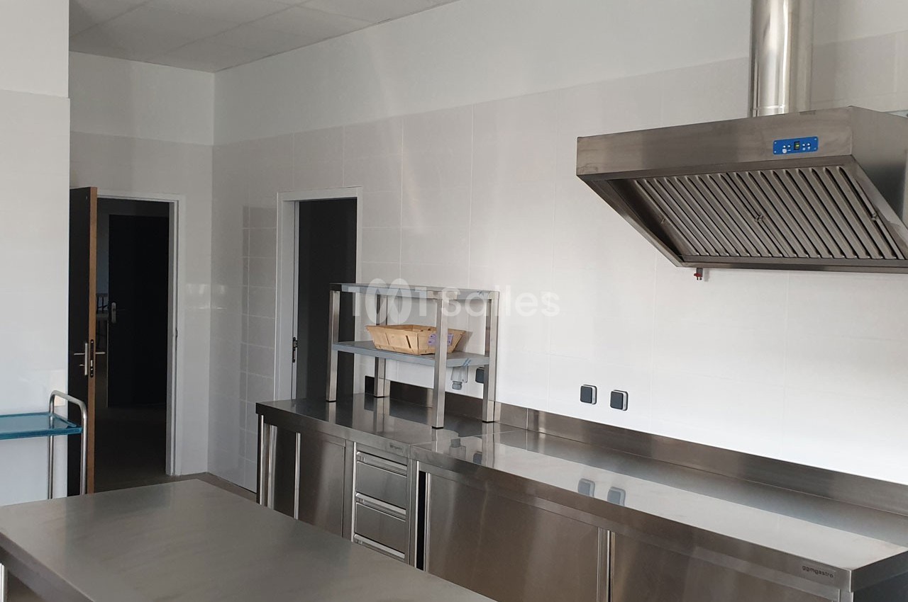Cuisine professionnelle avec plans de travail en inox, hotte aspirante, étagère métallique et murs carrelés blancs.