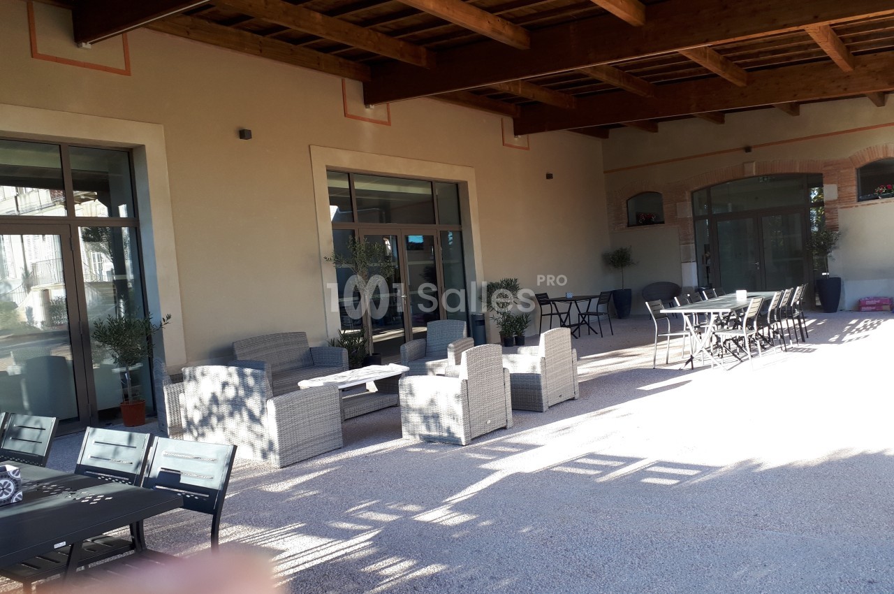 Terrasse couverte avec mobilier de jardin, tables et chaises, devant une façade avec grandes baies vitrées.
