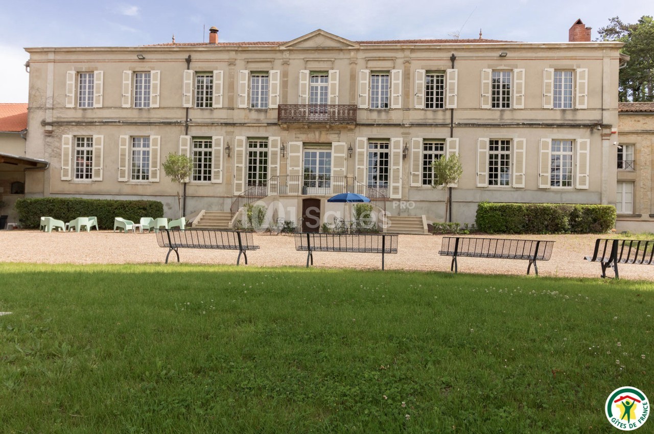 Façade d'un bâtiment ancien à deux étages avec des volets blancs, vue depuis une pelouse avec des bancs.