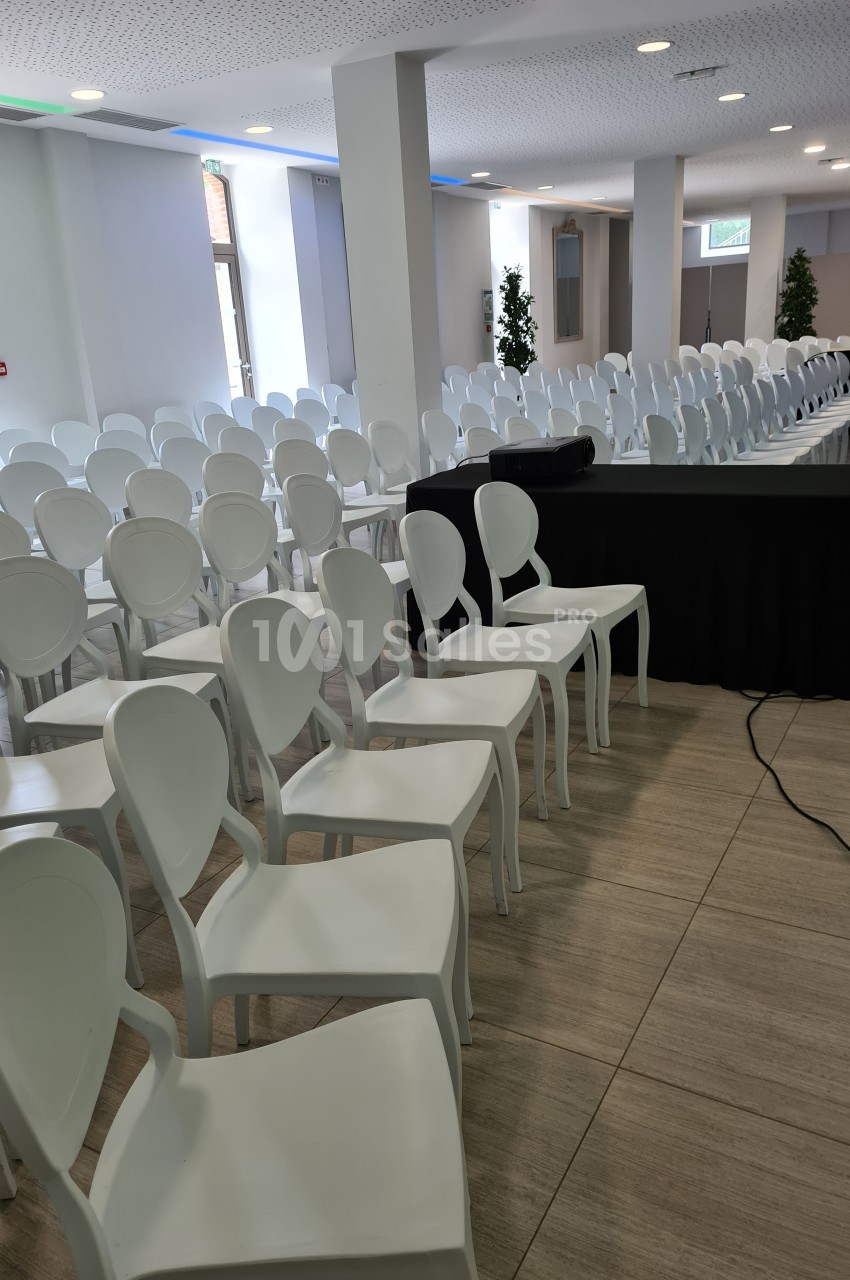 Salle de conférence lumineuse avec rangées de chaises blanches alignées et une table noire à l'avant.