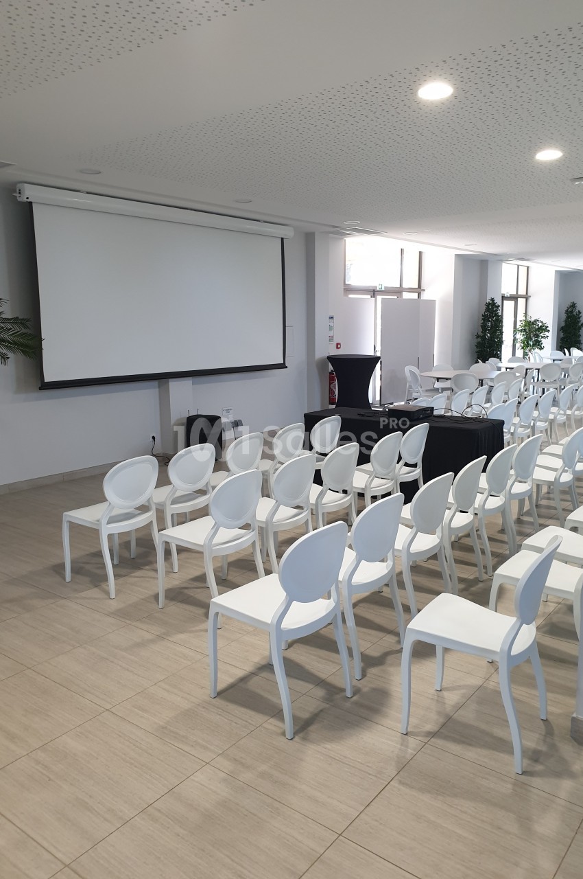 Salle de conférence lumineuse avec des rangées de chaises blanches alignées face à un écran de projection.