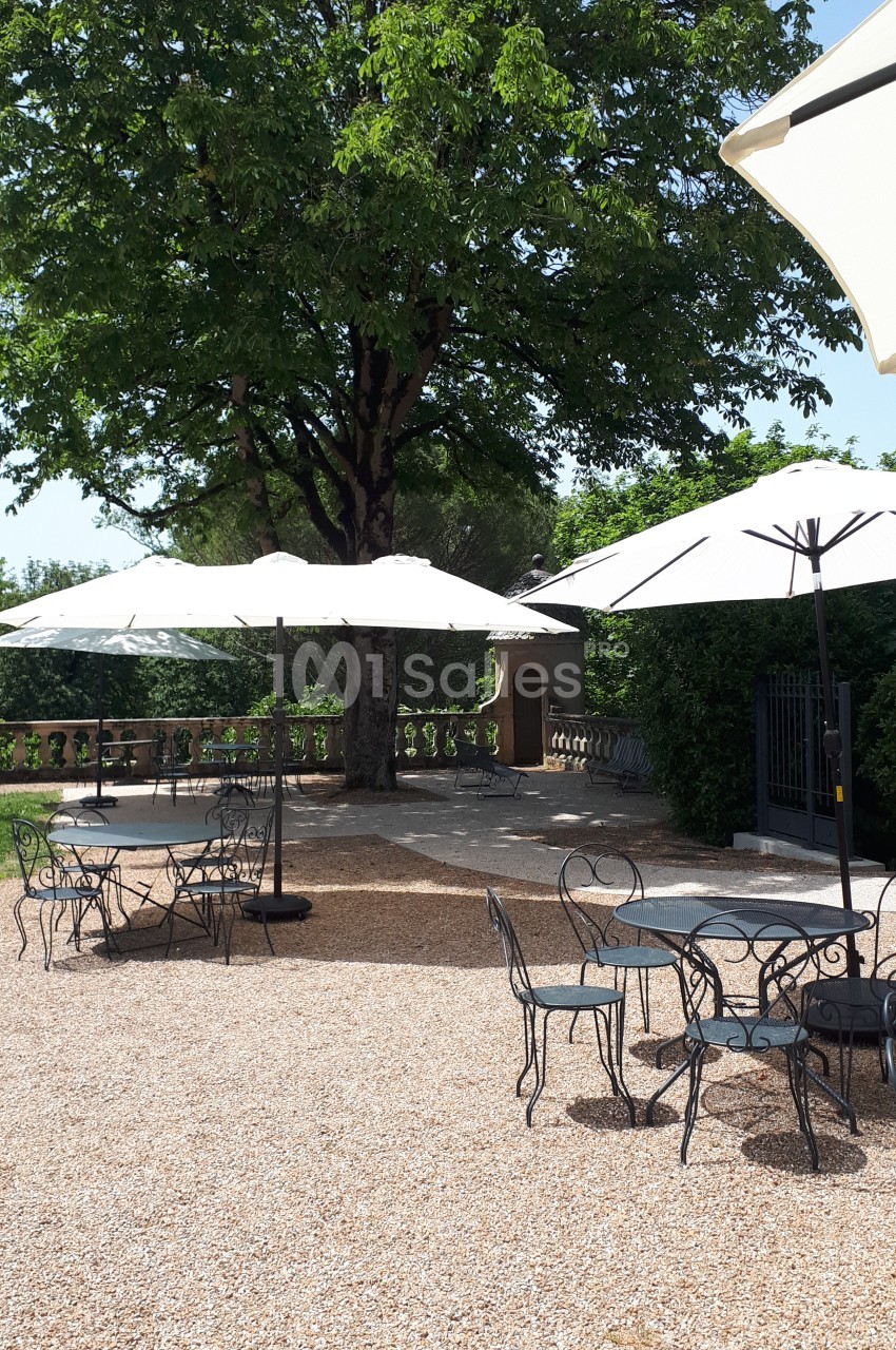 Terrasse en gravier avec tables et chaises en fer forgé, ombragée par des parasols et un grand arbre.