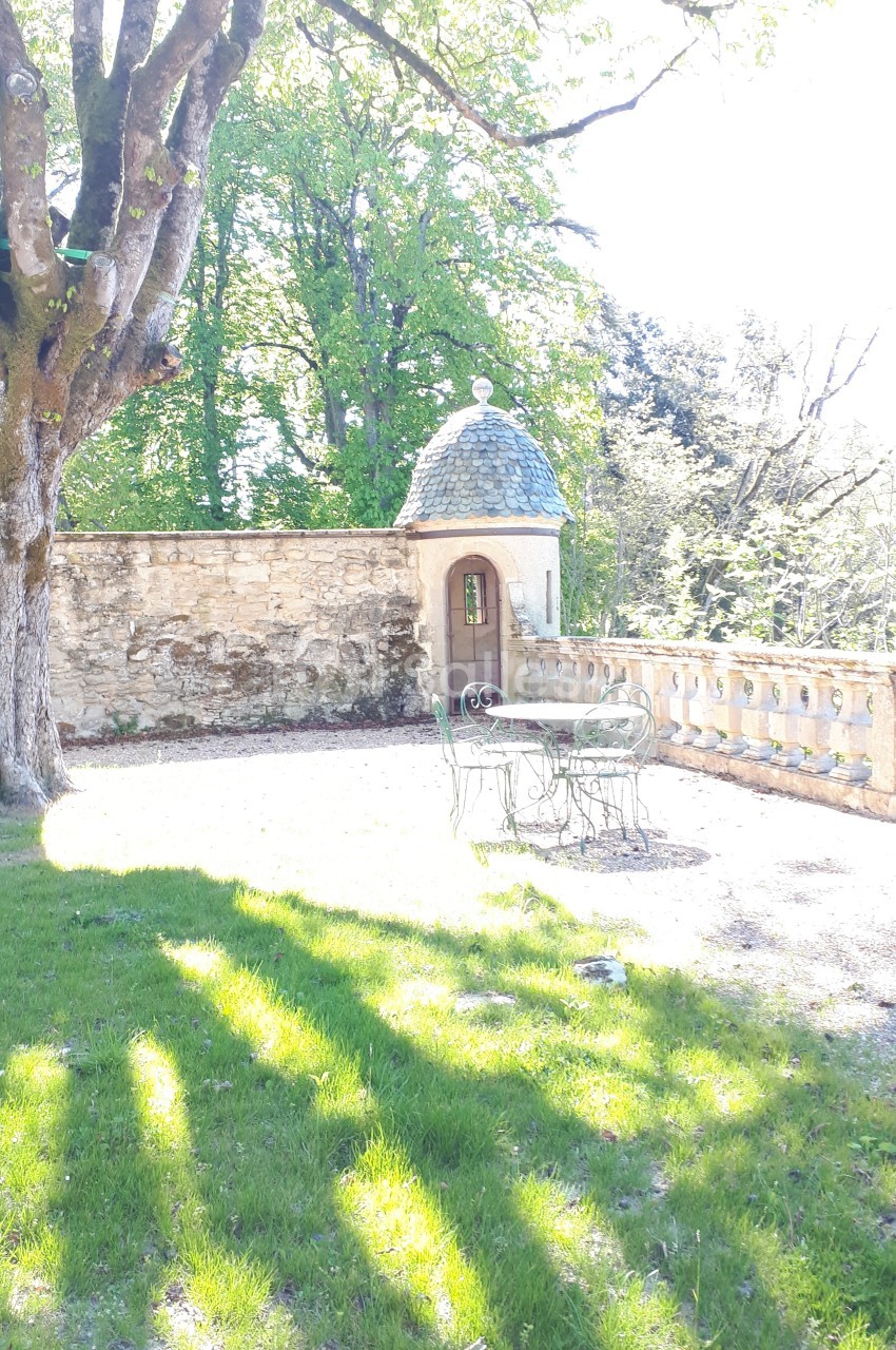 Terrasse ensoleillée avec table et chaises en fer forgé, bordée d'un mur en pierre et d'un petit pavillon.