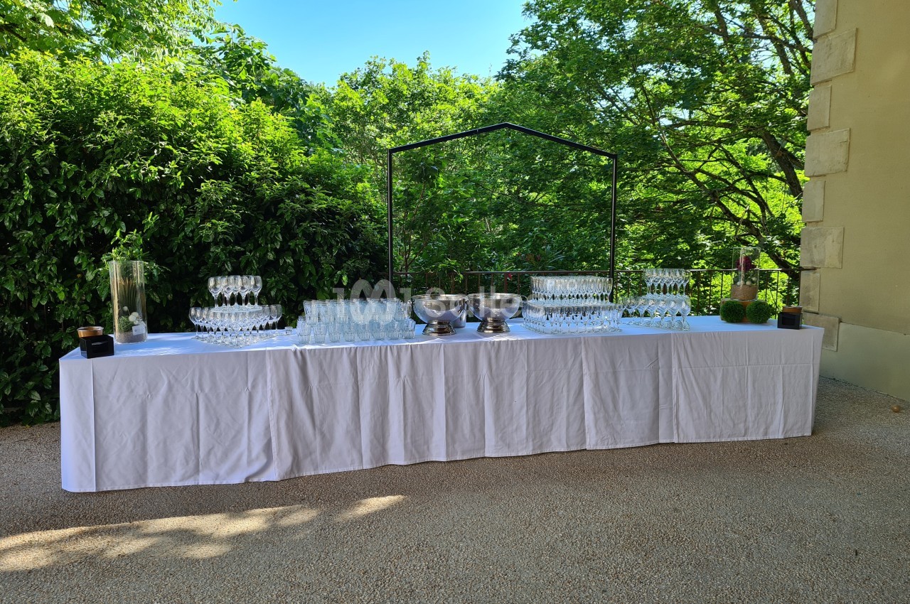 Table dressée en extérieur avec nappage blanc, verres à pied, seaux à champagne et verdure en arrière-plan.