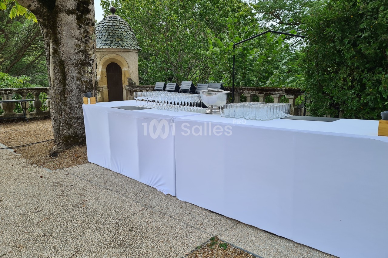 Table drapée de blanc avec des verres alignés et une vasque, installée en extérieur près d'une balustrade et d'un pavillon.