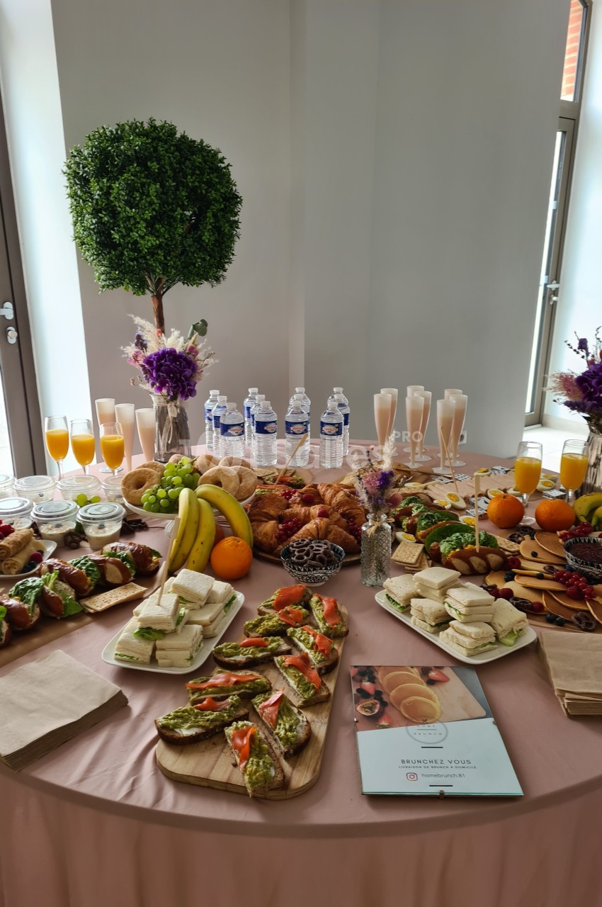 Table garnie de viennoiseries, fruits, sandwichs, boissons et décorations florales dans un cadre lumineux.