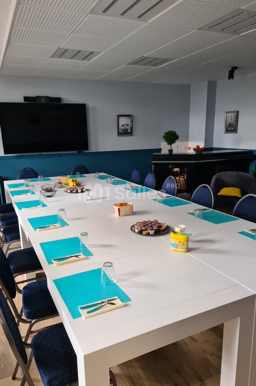 Salle de réunion avec une grande table blanche, sets de table bleus, chaises, écran mural et collations disposées.